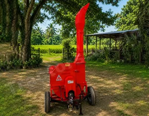 Red portable wood chipper on wheels in a rural yard, with a tall curved discharge chute and green fields in the background.