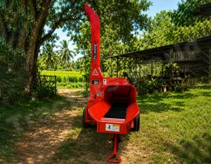 Red wood chipper on a trailer in a grassy rural yard, with a dirt path and trees in the background